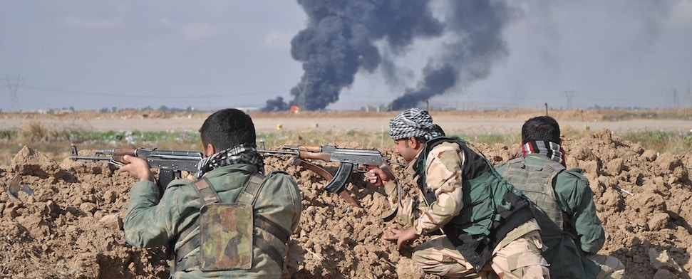 Mehrere Peschmerga Kämpfer liegen hinter einem Erdwall nahe der irakischen Stadt Kirkuk. Im Hintergrund sind Rauchsäulen zu sehen.
